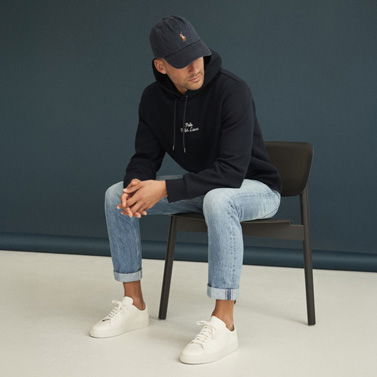 Mann sitzt auf Stuhl, trägt dunkelblauen Hoodie, hellblaue Jeans, weiße Sneaker und dunkelblaue Cap, Blick nach unten. Mann sitzt auf Stuhl, trägt dunkelblauen Hoodie, hellblaue Jeans, weiße Sneaker und dunkelblaue Cap, Blick nach unten.