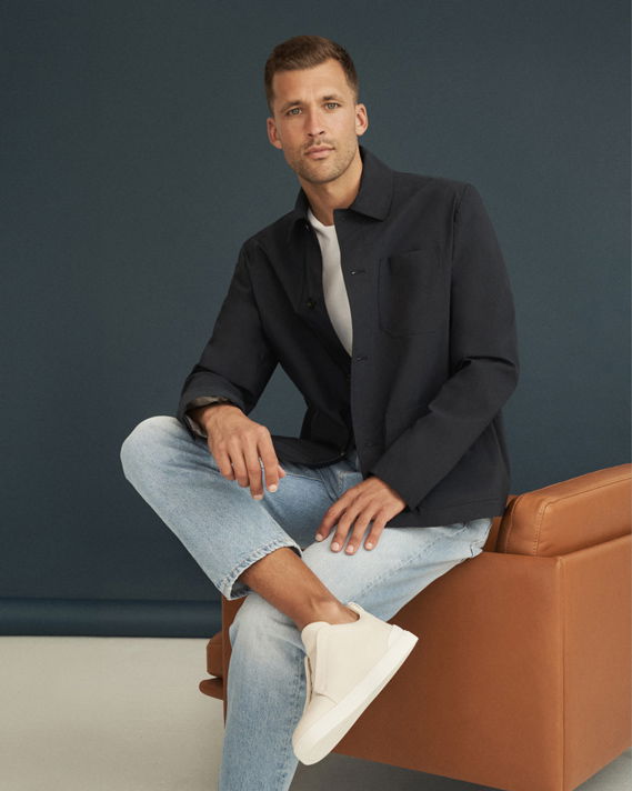 Mann sitzt auf braunem Sessel, trägt hellblaue Jeans, weißes Shirt, dunkelblaues Overshirt und weiße Sneaker, vor dunklem Hintergrund. Mann sitzt auf braunem Sessel, trägt hellblaue Jeans, weißes Shirt, dunkelblaues Overshirt und weiße Sneaker, vor dunklem Hintergrund.