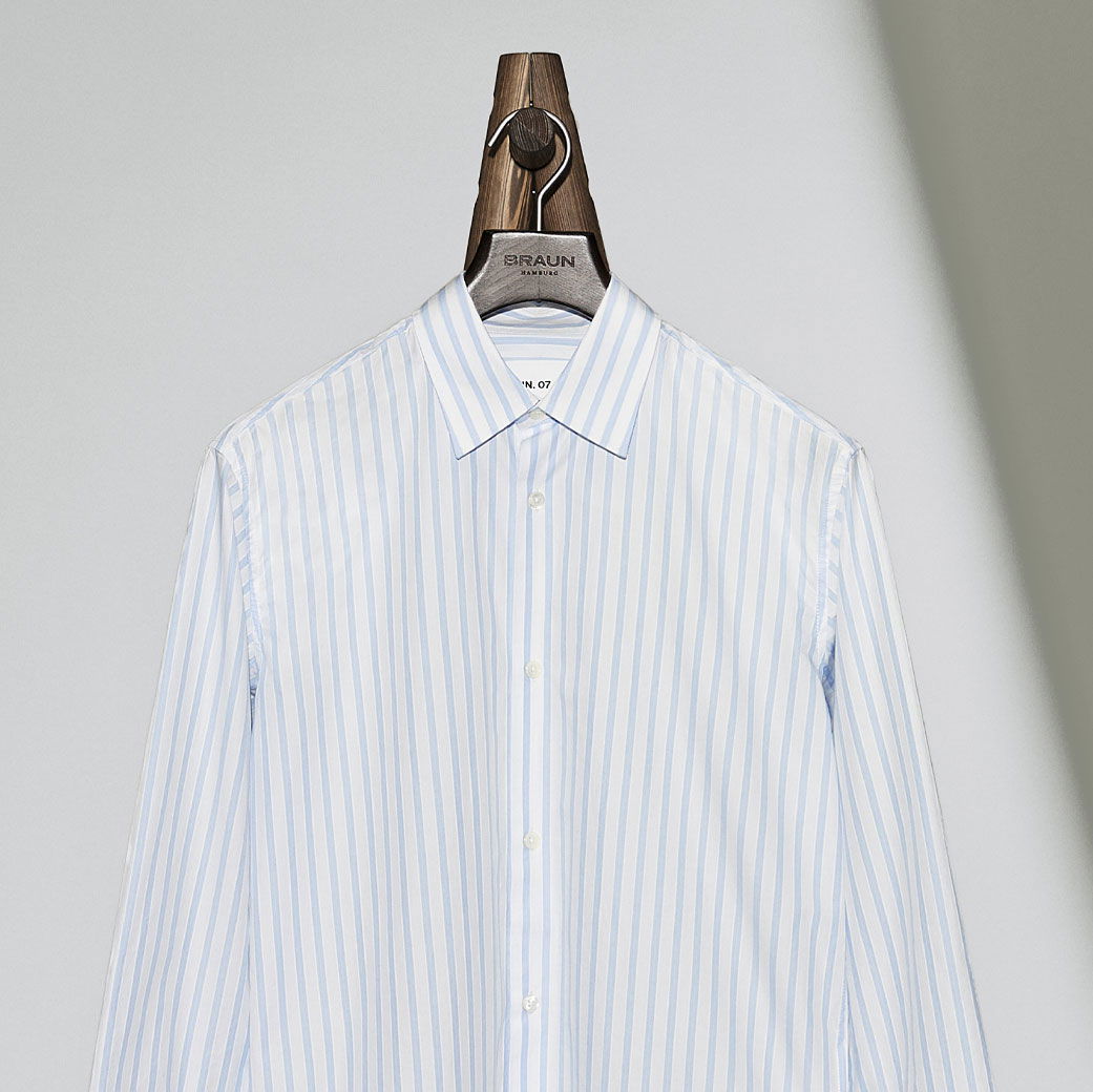 Hellblau-weiß gestreiftes Herrenhemd mit Knöpfen, auf dunkelbraunem Holzbügel vor hellgrauem Hintergrund.