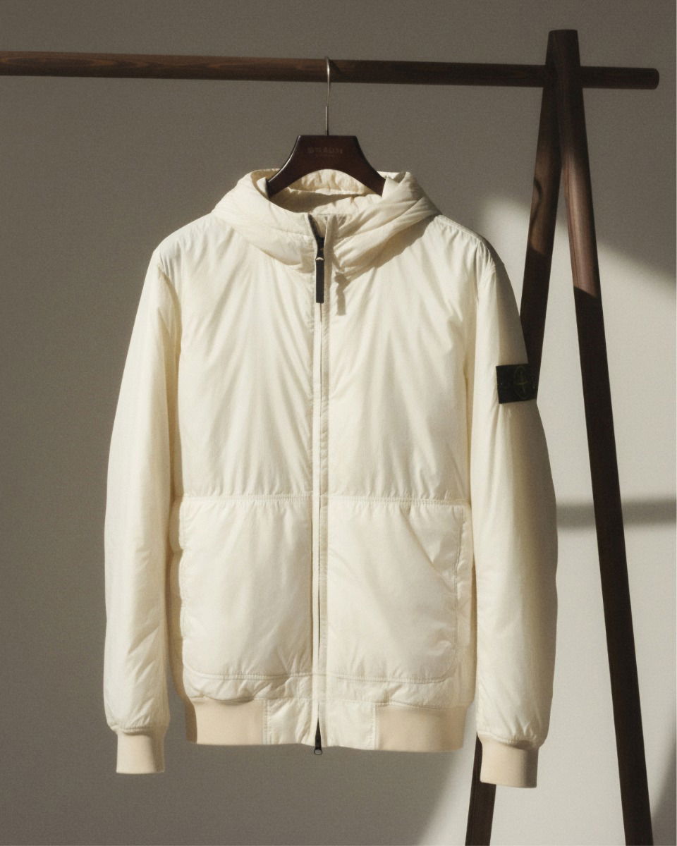 Leichte, cremefarbene Kapuzenjacke mit Zwei-Wege-Reißverschluss und Stone Island Logo-Patch am linken Ärmel, hängend. Leichte, cremefarbene Kapuzenjacke mit Zwei-Wege-Reißverschluss und Stone Island Logo-Patch am linken Ärmel, hängend.