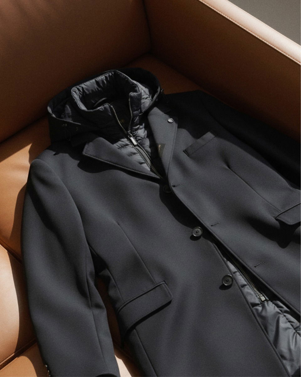 Schwarzer Mantel mit Steppjacke und Kapuze liegt zerknittert auf braunem Ledersofa in hellem Licht. Schwarzer Mantel mit Steppjacke und Kapuze liegt zerknittert auf braunem Ledersofa in hellem Licht.