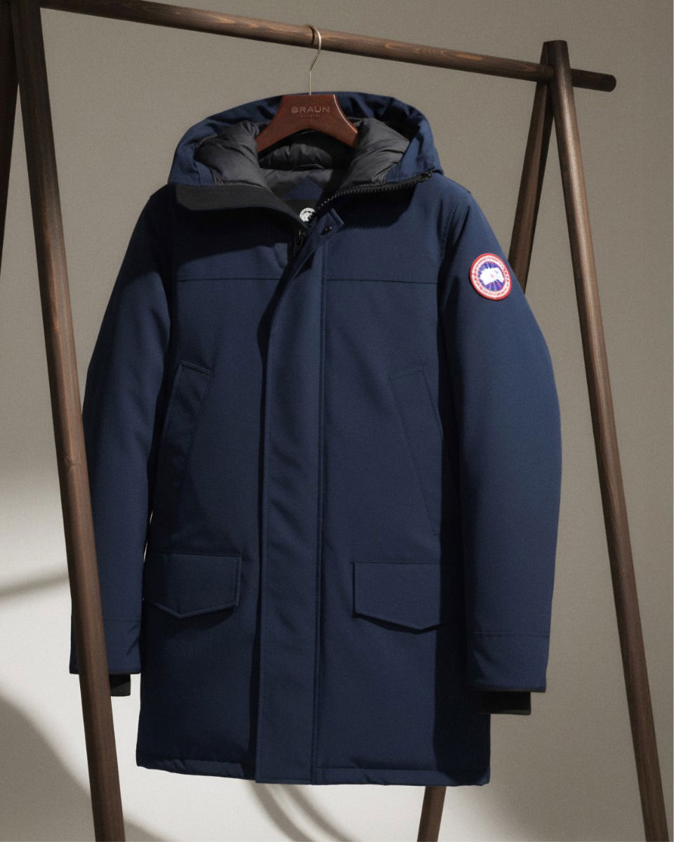 Dunkelblauer Winterparka mit Kapuze, Reißverschluss und Taschen, hängt an einem Holzkleiderständer vor neutralem Hintergrund. Dunkelblauer Winterparka mit Kapuze, Reißverschluss und Taschen, hängt an einem Holzkleiderständer vor neutralem Hintergrund.