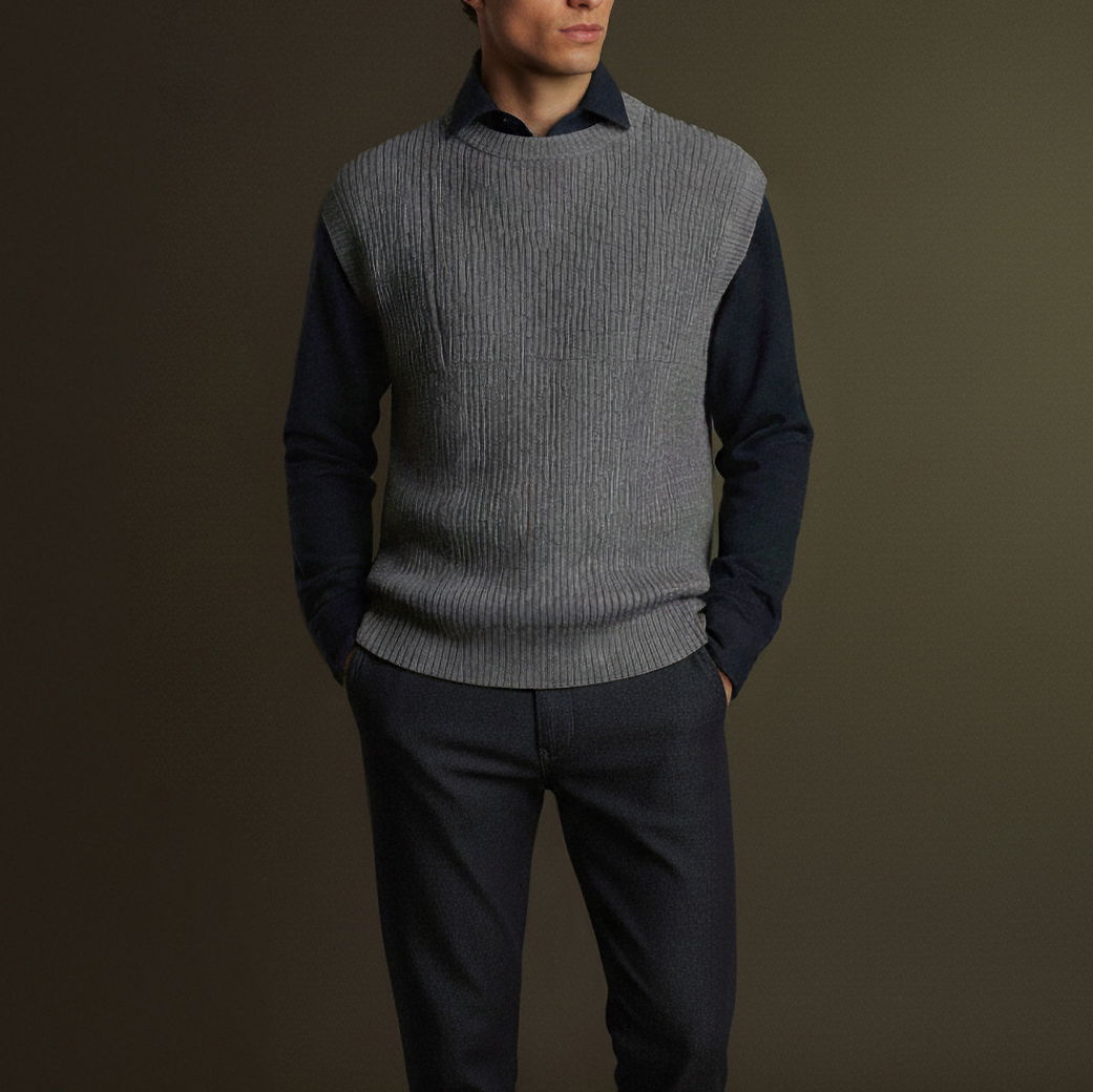 Mann trägt dunkelblaues Hemd, graues Strickweste ohne Ärmel und dunkle Hose, steht vor schlichtem Hintergrund.