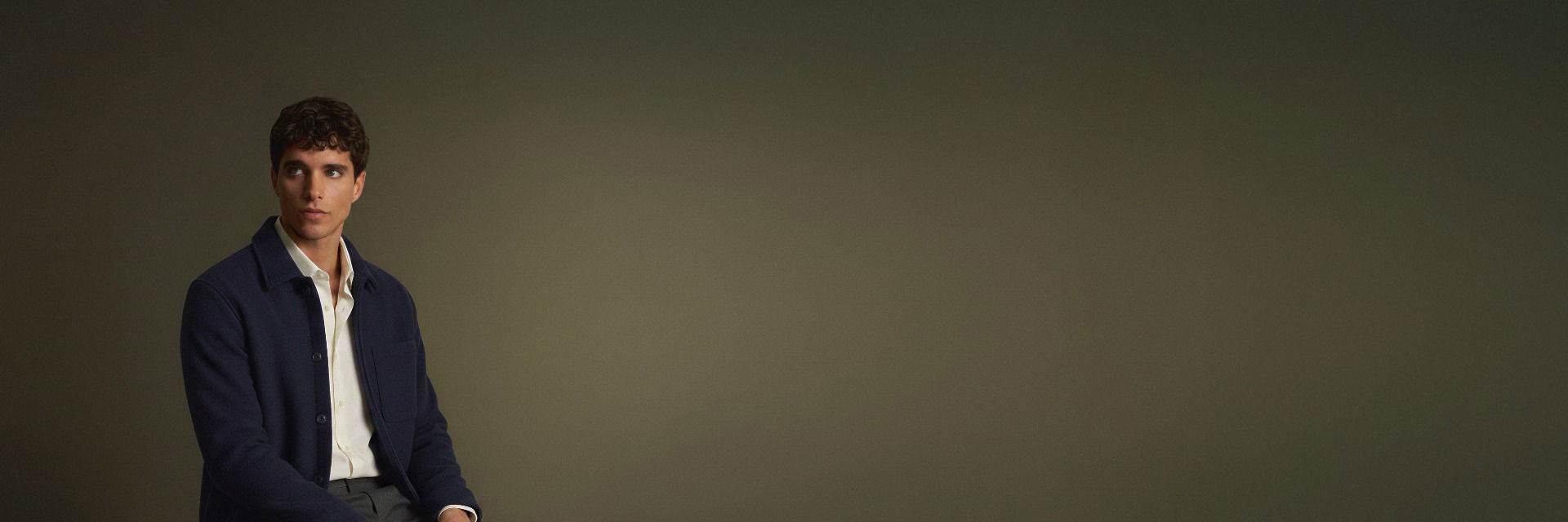 Junger Mann mit kurzen lockigen Haaren, trägt weiße Hemd und dunkelblaue Jacke, vor schlichtem braunem Hintergrund. Junger Mann mit kurzen lockigen Haaren, trägt weiße Hemd und dunkelblaue Jacke, vor schlichtem braunem Hintergrund.