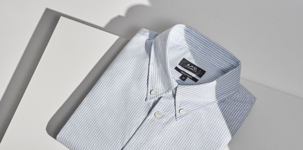 Gefaltetes Herrenhemd mit feinen blauen Streifen und Knöpfen auf weißem Tisch, von oben fotografiert.