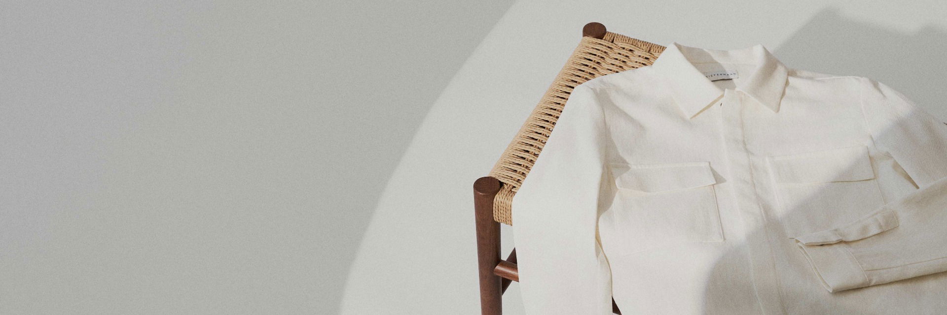 Weißes Herrenhemd mit Kragen und Brusttaschen liegt von oben fotografiert auf einem Holzstuhl mit geflochtenem Sitz. Weißes Herrenhemd mit Kragen und Brusttaschen liegt von oben fotografiert auf einem Holzstuhl mit geflochtenem Sitz.