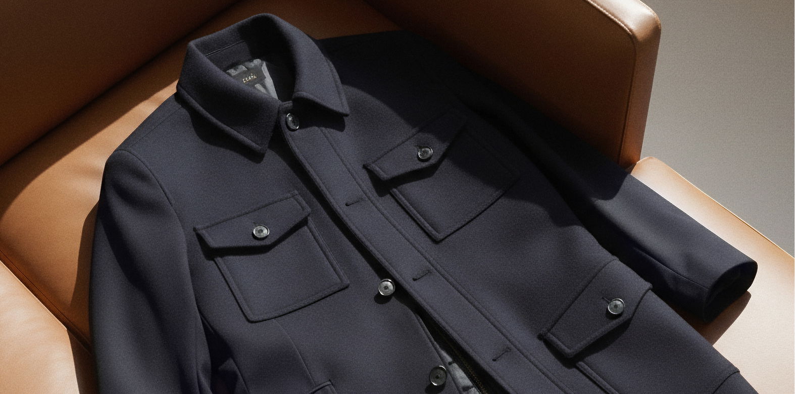 Dunkelblaue Jacke mit Kragen und vier aufgesetzten Taschen liegt auf braunem Ledersessel im Sonnenlicht. Dunkelblaue Jacke mit Kragen und vier aufgesetzten Taschen liegt auf braunem Ledersessel im Sonnenlicht.