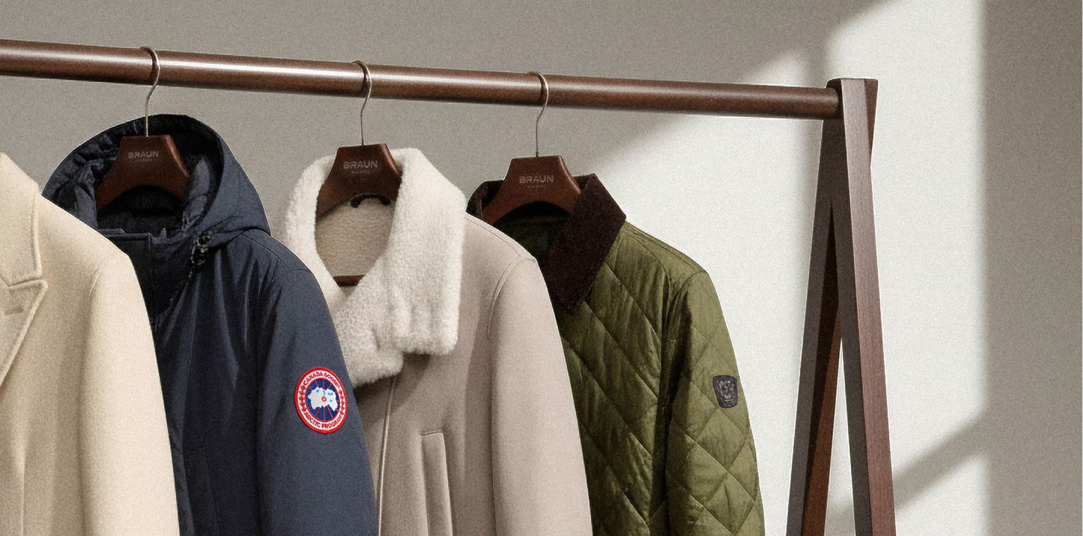 Vier Jacken in Beige, Dunkelblau mit Logo, Hellbeige mit Fellkragen und Grün mit Steppmuster hängen an einem Holz-Kleiderständer. Vier Jacken in Beige, Dunkelblau mit Logo, Hellbeige mit Fellkragen und Grün mit Steppmuster hängen an einem Holz-Kleiderständer.