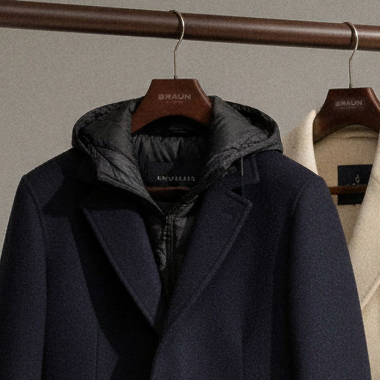 Dunkelblauer Mantel mit Stepp-Innenseite und Kapuze sowie cremefarbener Mantel auf hölzernen Braun-Kleiderbügeln an Metallstange. Dunkelblauer Mantel mit Stepp-Innenseite und Kapuze sowie cremefarbener Mantel auf hölzernen Braun-Kleiderbügeln an Metallstange.