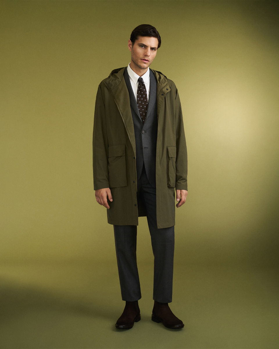 Mann trägt grauen Anzug mit weißem Hemd, brauner gepunkteter Krawatte, olivgrünem Mantel und braunen Stiefeln vor grünem Hintergrund. Mann trägt grauen Anzug mit weißem Hemd, brauner gepunkteter Krawatte, olivgrünem Mantel und braunen Stiefeln vor grünem Hintergrund.