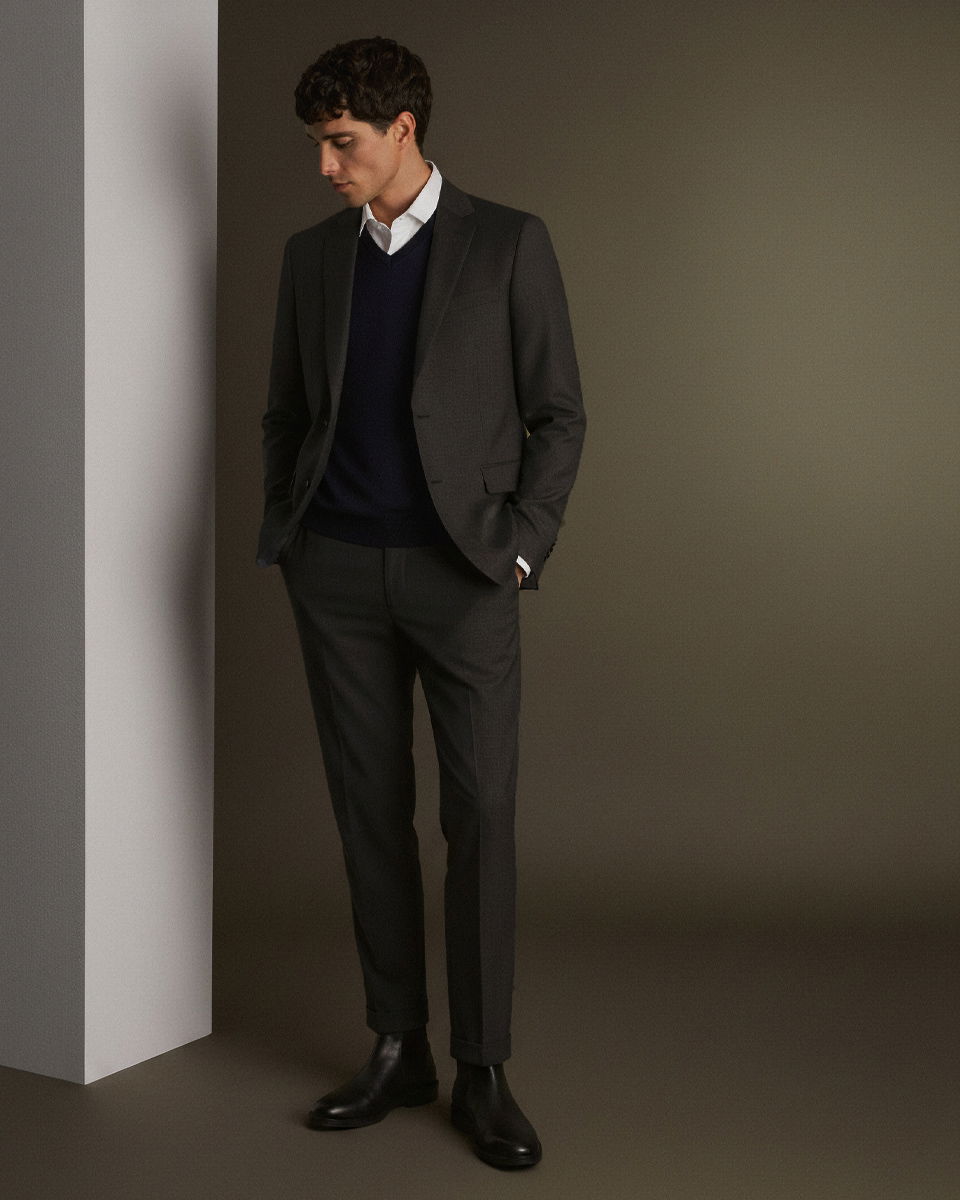 Junger Mann im dunkelgrauen Anzug, weißem Hemd und schwarzem Pullover, steht mit Händen in Taschen vor grauem Hintergrund.
