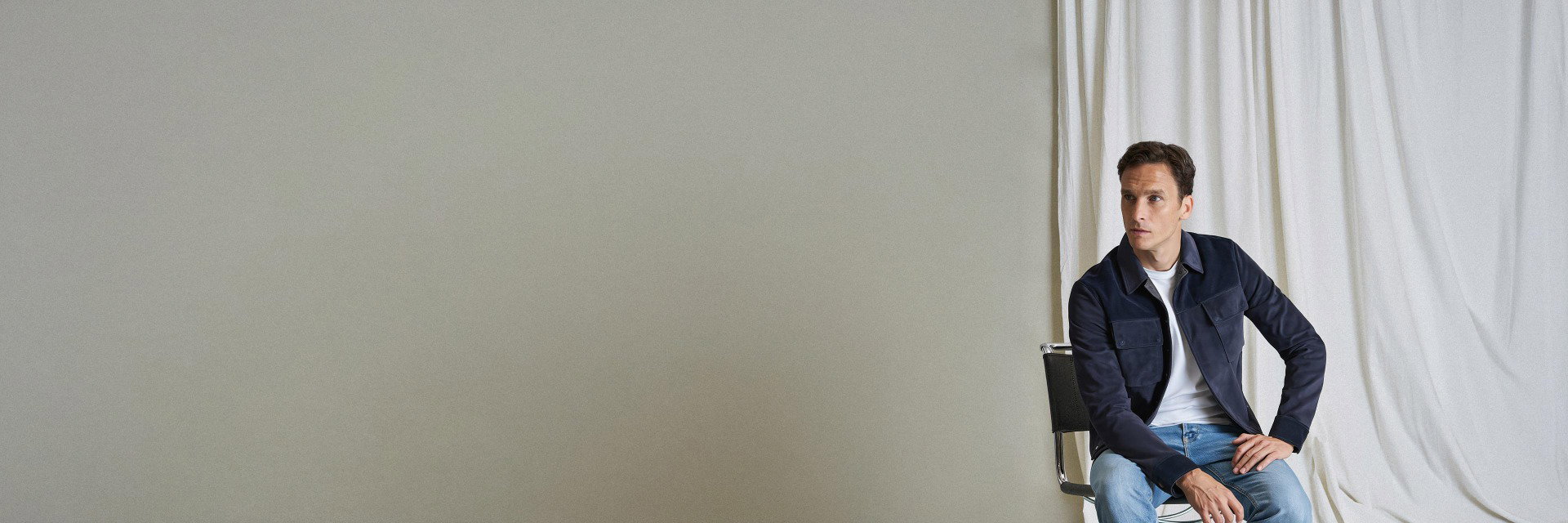 Mann sitzt auf Stuhl vor grauer Wand und weißem Vorhang, trägt blaue Jacke, weißes T-Shirt und blaue Jeans. Mann sitzt auf Stuhl vor grauer Wand und weißem Vorhang, trägt blaue Jacke, weißes T-Shirt und blaue Jeans.