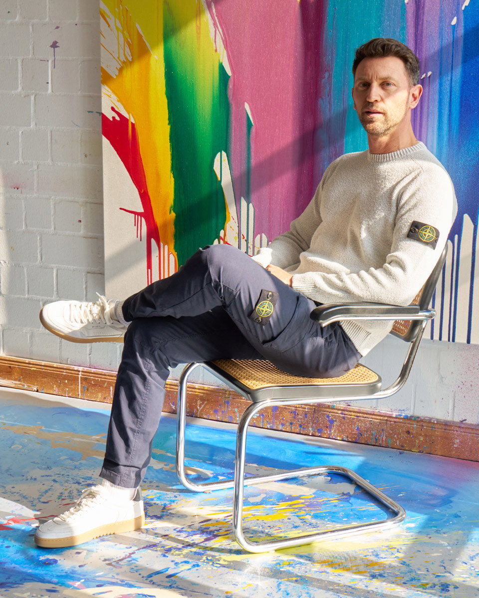 Mann sitzt auf Stuhl mit Flechtgeflecht, trägt hellen Pullover, dunkle Hose, weiße Sneaker, bunte Farbkleckse an Wand und Boden.