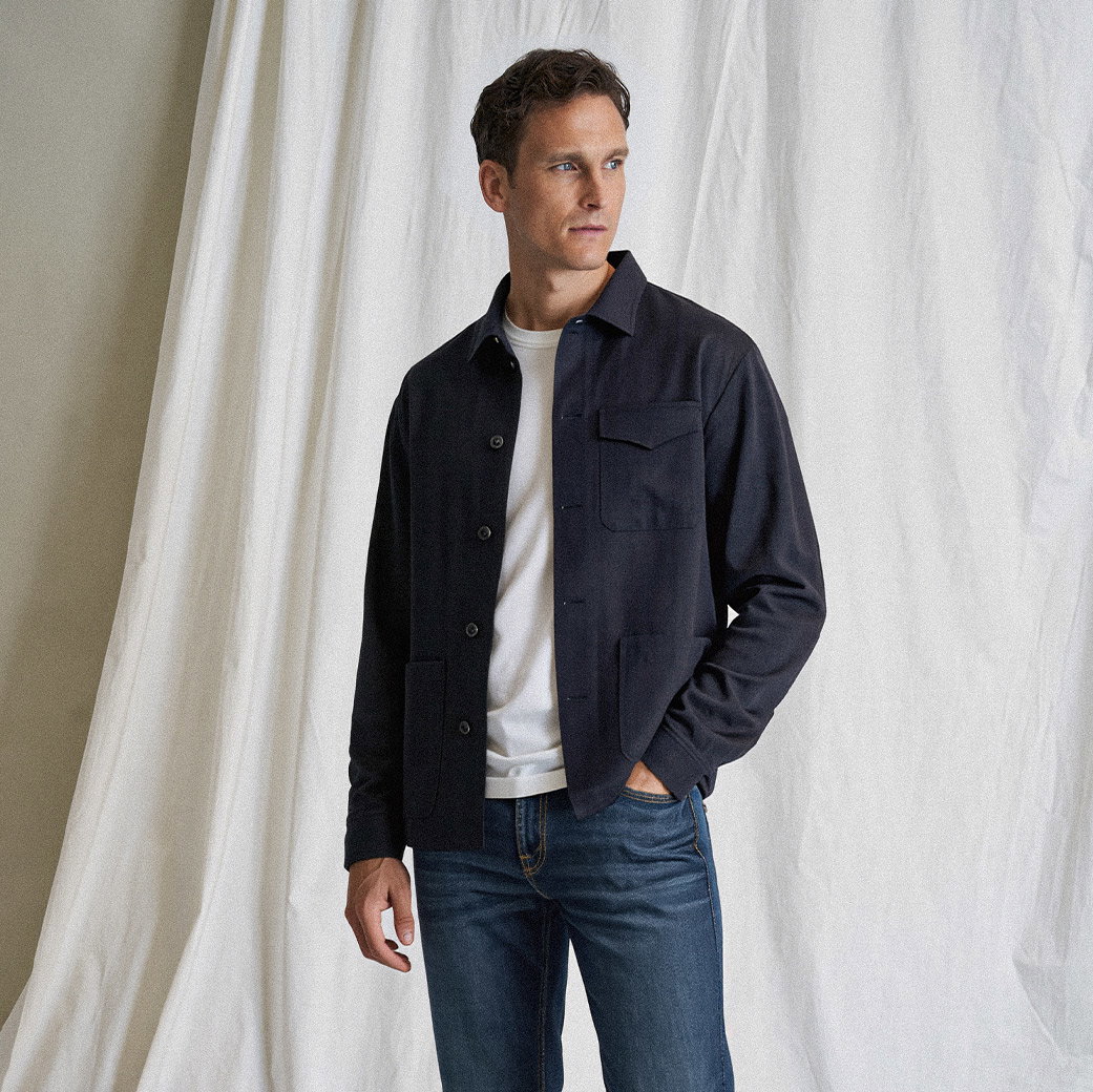 Mann mit blauen Jeans, weißem T-Shirt und dunkelblauer Jacke steht vor weißem Vorhang und schaut seitlich.