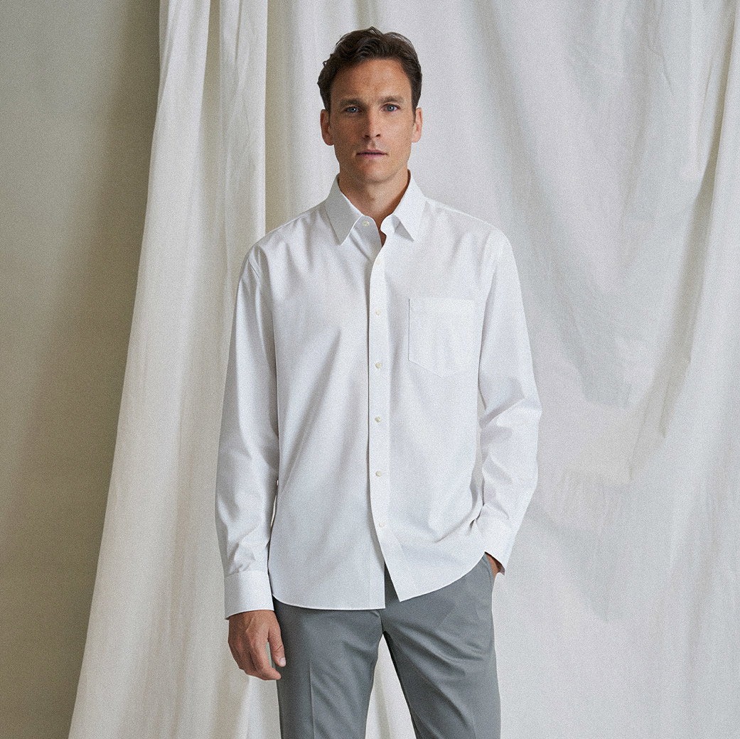 Mann mit kurzen braunen Haaren trägt weiße Hemdjacke mit Brusttasche und graue Hose, steht vor hellem Stoffhintergrund.