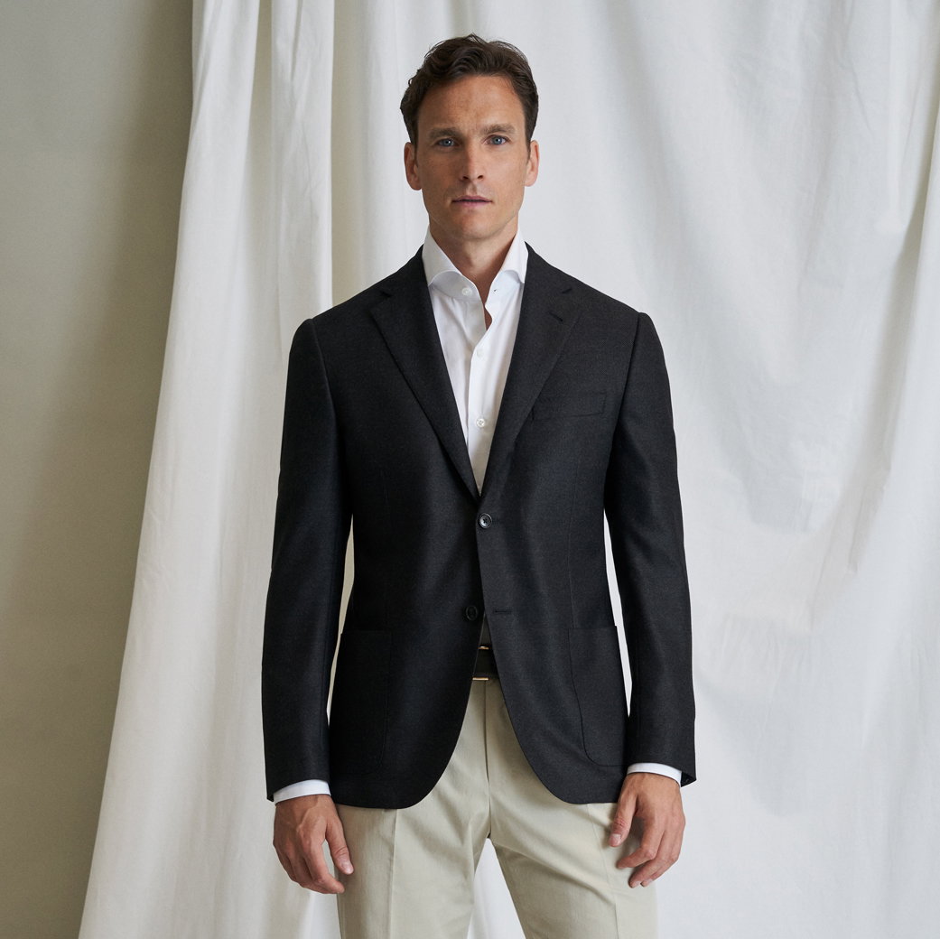 Mann mit kurzem braunem Haar trägt schwarzen Blazer, weißes Hemd und beige Hose, steht vor weißem Stoffhintergrund.