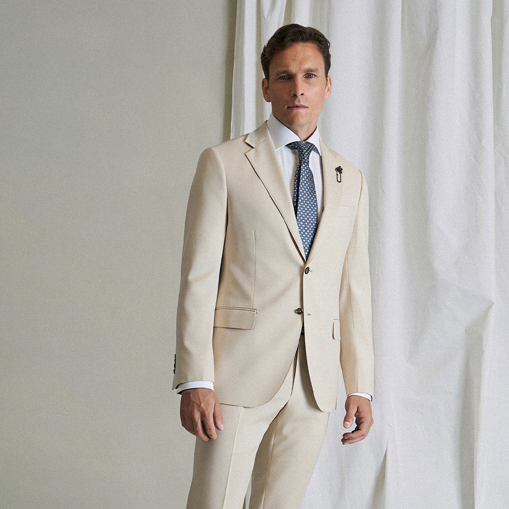 Mann trägt eleganten hellbeigen Anzug mit weißem Hemd und gemustertem blauen Krawatte vor hellem Vorhang.
