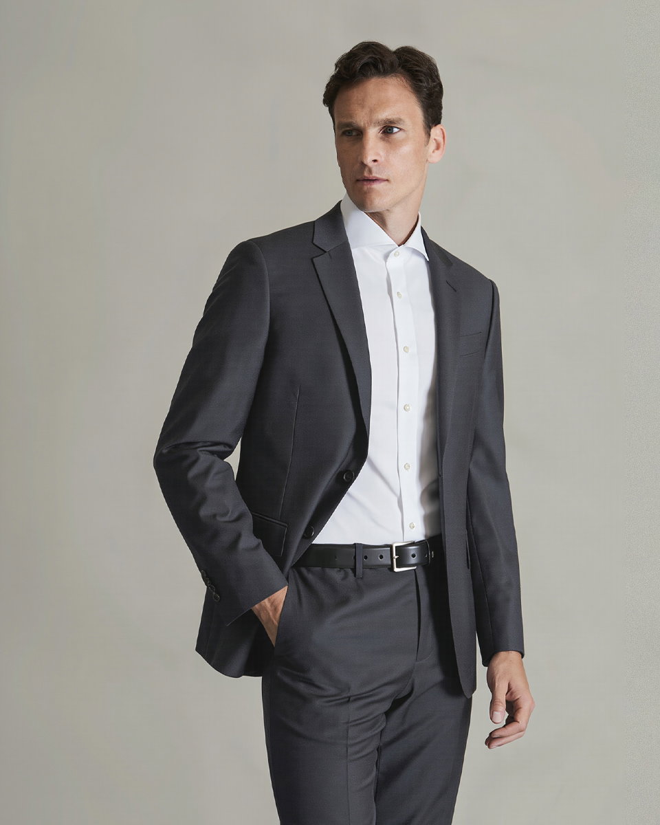 Mann im schwarzen Anzug und weißem Hemd, mit linker Hand in Hosentasche, schaut nach rechts vor hellem Hintergrund.