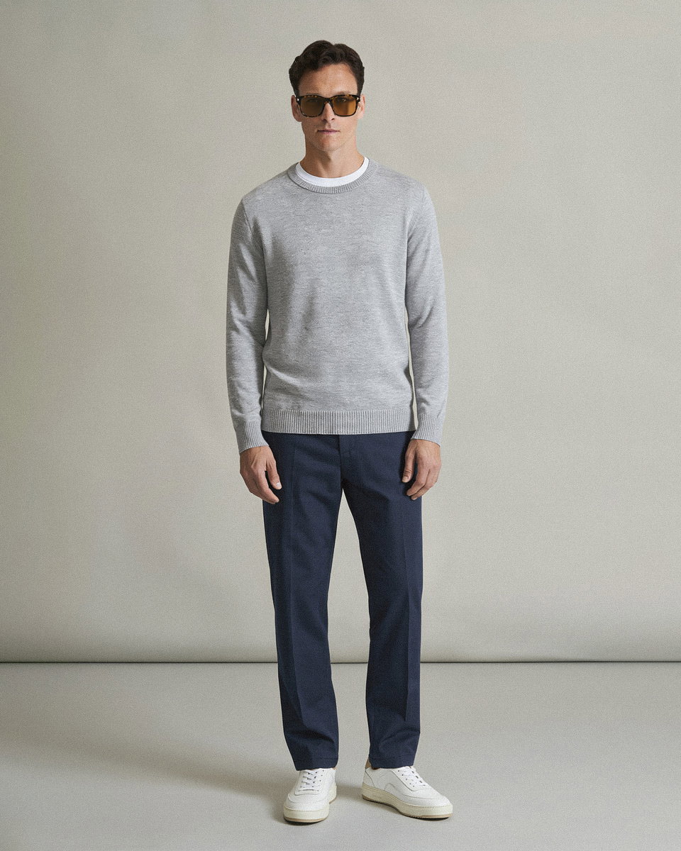 Mann trägt grauen Pullover, weiße Sneakers, dunkelblaue Hose und braune Sonnenbrille, steht vor neutralem Hintergrund.