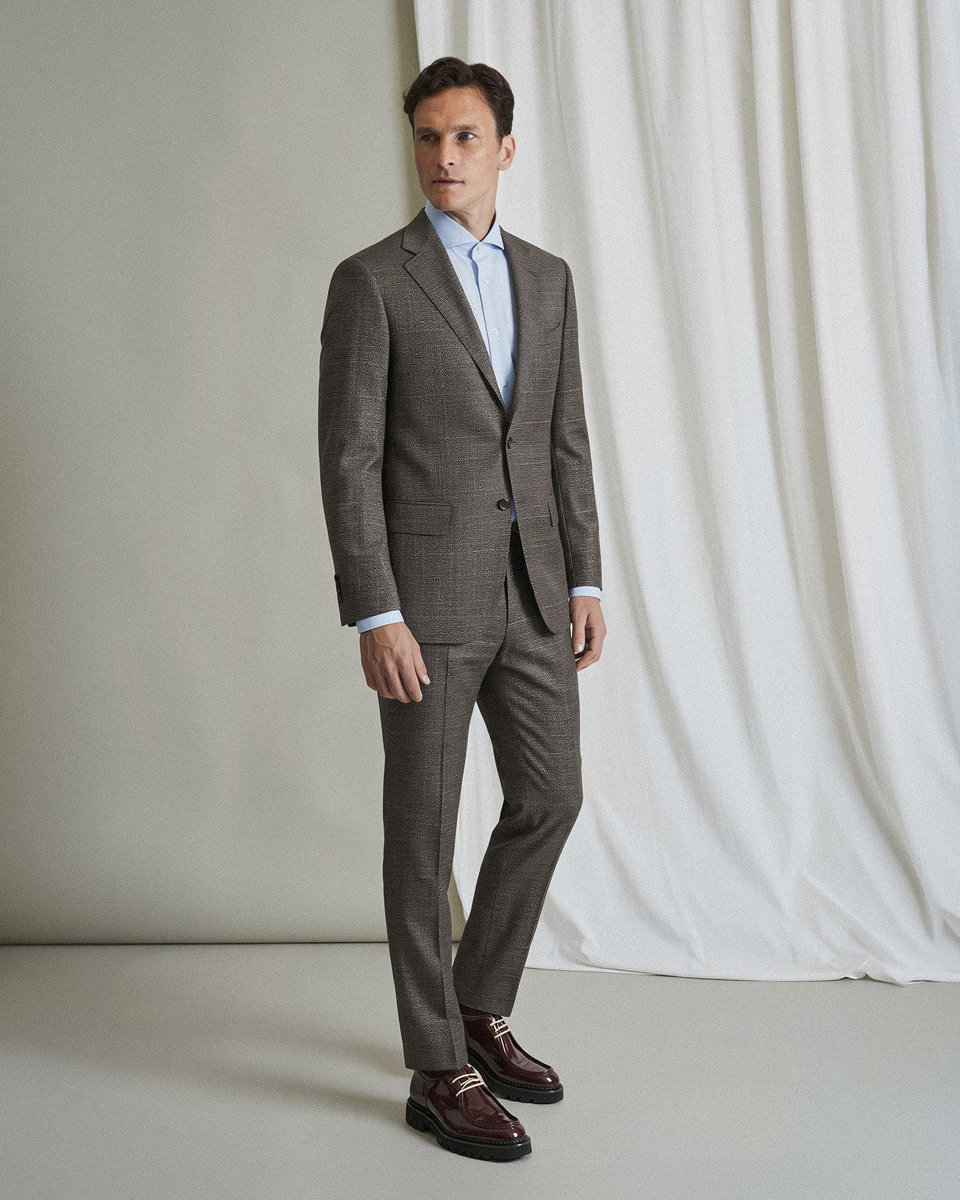 Mann trägt braunen Anzug mit hellblauem Hemd und dunkelroten Schnürschuhen, steht vor weißem Vorhang.