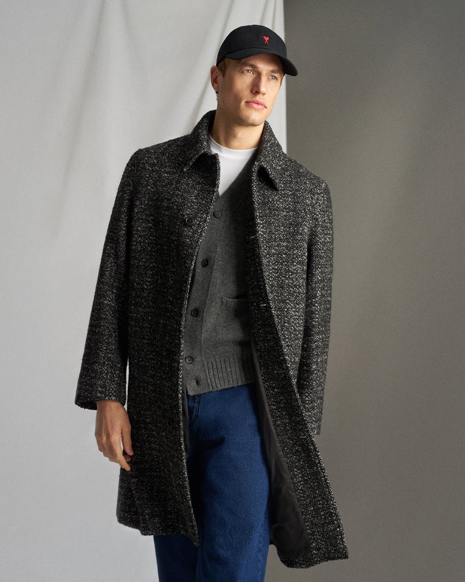 Mann trägt schwarzen Cap, grauen Cardigan, weißen Pullover und schwarzen Wollmantel, dazu blaue Jeans vor grauem Hintergrund.