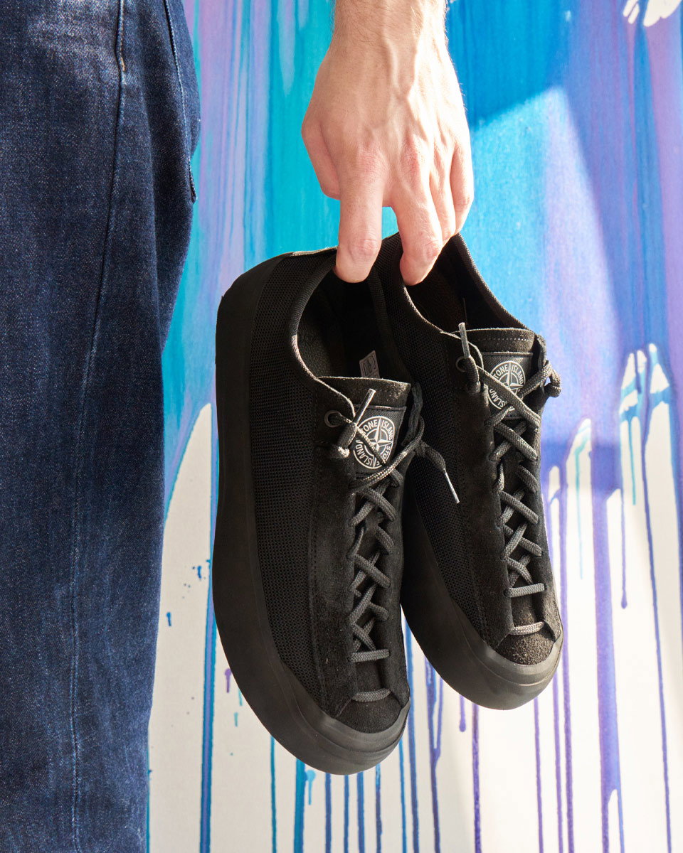 Hand hält ein Paar schwarze Stone Island Sneaker vor bunter Wand mit blauen und lila Farbtropfen, Jeans sichtbar.