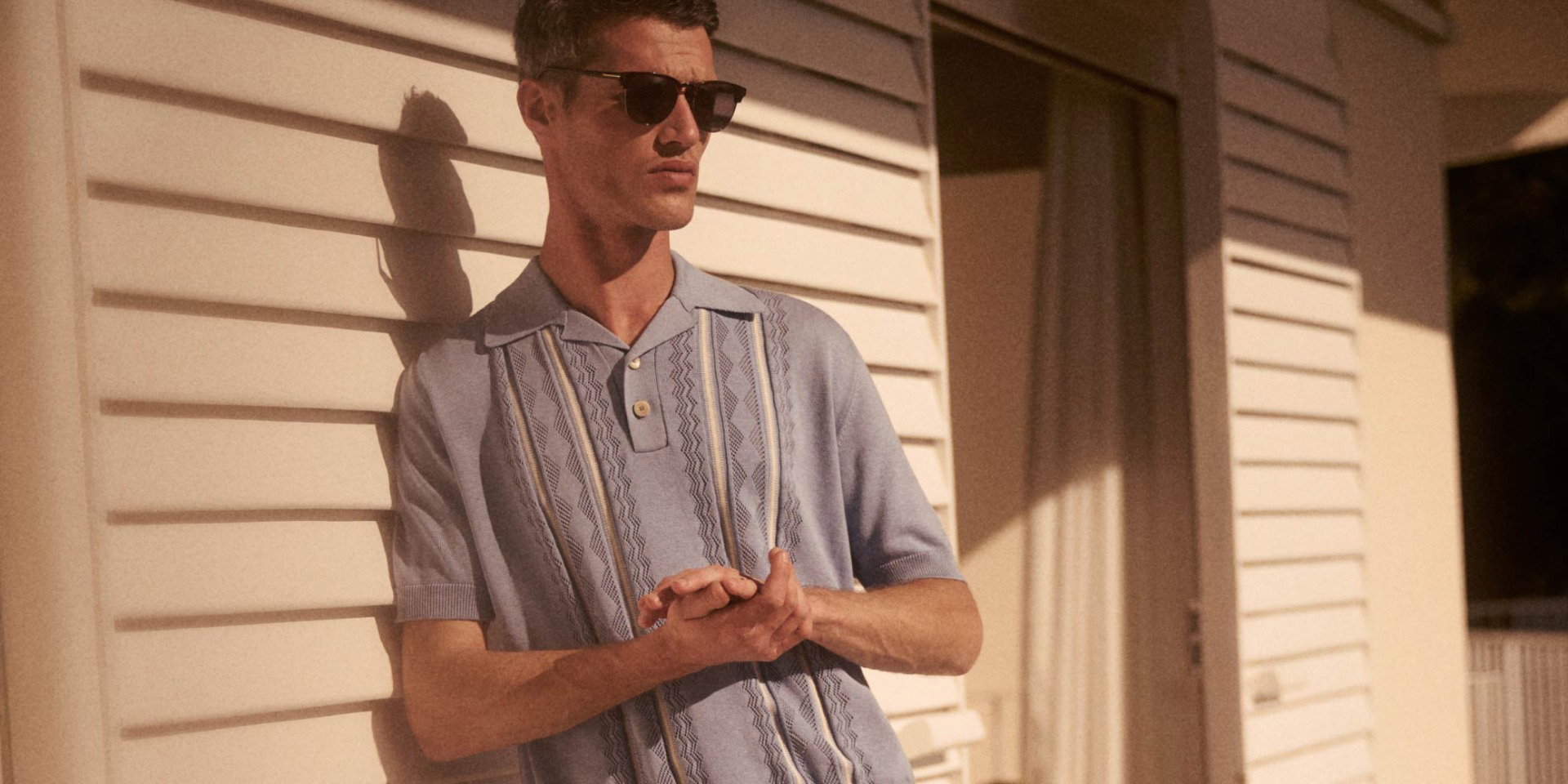 Junger Mann mit Sonnenbrille lehnt an weißer Holzfassade, trägt hellblaues gemustertes Kurzarmhemd, sonniger Tag.