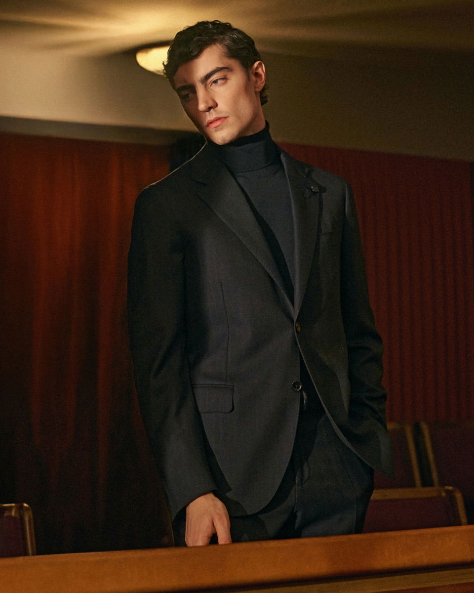 Junger Mann mit dunklem Anzug und Rollkragenpullover steht nachdenklich in einem gedämpft beleuchteten Raum mit Holzvertäfelung.