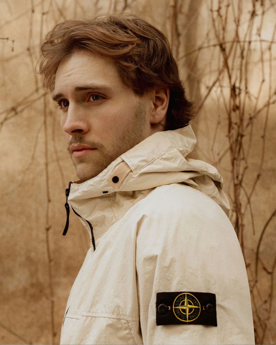 Porträt eines jungen Mannes mit braunen Haaren in heller Stone Island Jacke vor beiger Wand mit Kletterpflanzen.