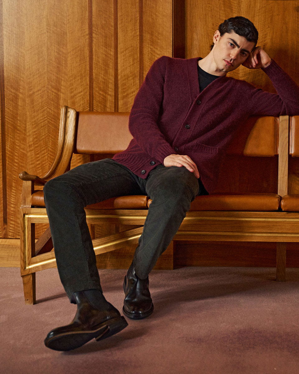 Junger Mann lehnt entspannt auf braunem Ledersofa, trägt bordeauxroten Strickcardigan, schwarze Hose und schwarze Stiefel.