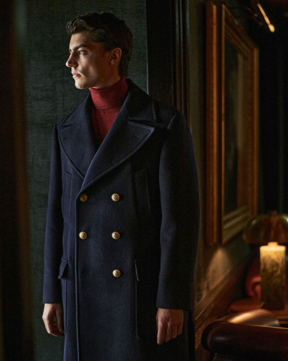 Junger Mann im dunkelblauen, zweireihigen Mantel mit goldenen Knöpfen und rotem Rollkragenpullover, stehend in elegantem Raum.