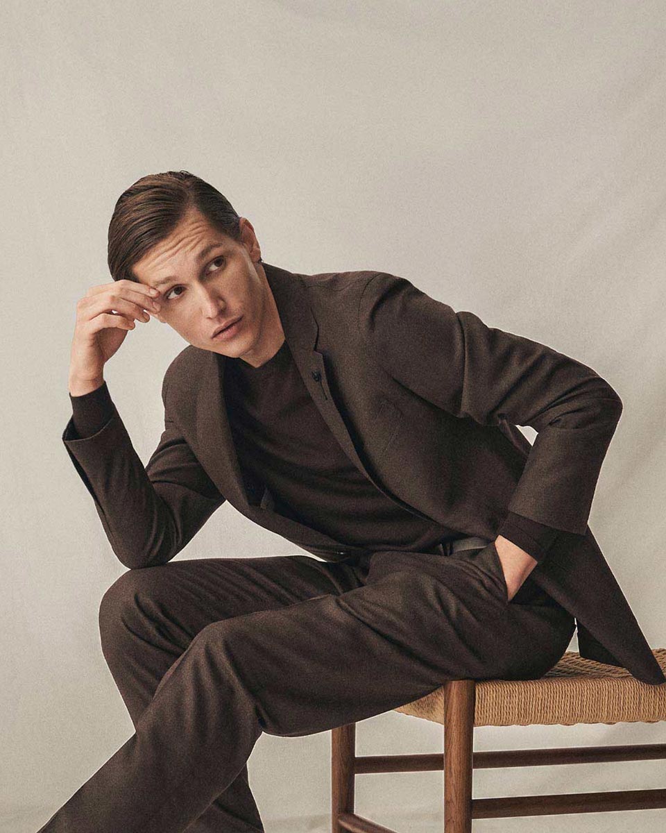 Junger Mann mit braunem Anzug und schwarzem Pullover sitzt nachdenklich auf einem Holzstuhl vor hellem Hintergrund.