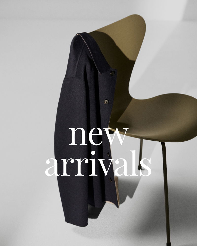 Dunkelblauer Mantel hängt über die Rückenlehne eines olivgrünen Stuhls vor weißem Hintergrund mit Text 'new arrivals'.
