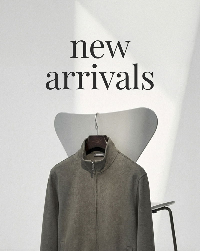 Graue Reißverschlussjacke an einem Holzkleiderbügel vor einem weißen Stuhl mit Schriftzug „new arrivals“ im Hintergrund.