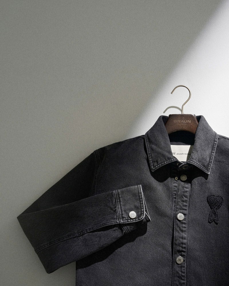 Schwarze Jeansjacke mit Herz-Logo links auf Brust, auf Holzbügel mit 'Braun'-Prägung, im Licht vor grauer Wand hängend.