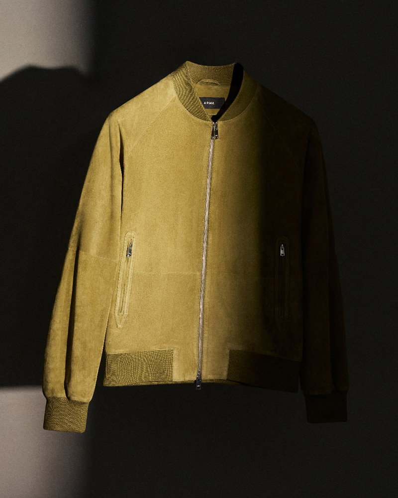 Olive green suede bomber jacket with ribbed collar, cuffs, hem, and front zipper pockets displayed against a dark background.