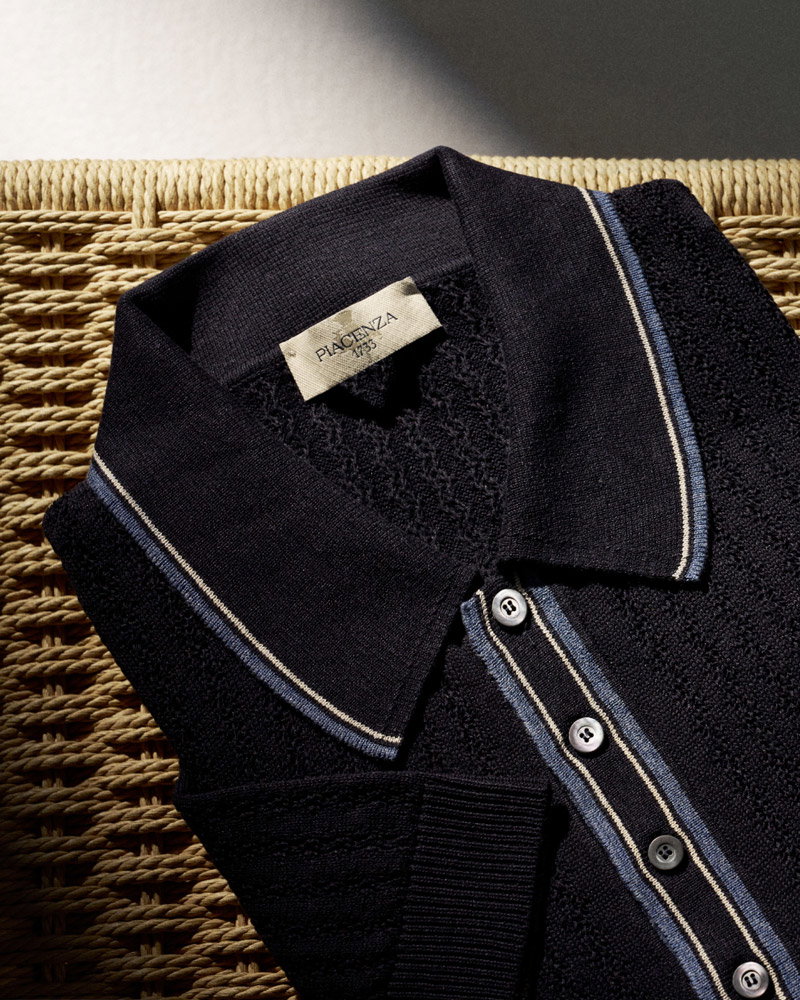 Folded black textured knit polo shirt with blue and white striped collar and button placket, placed on woven surface.