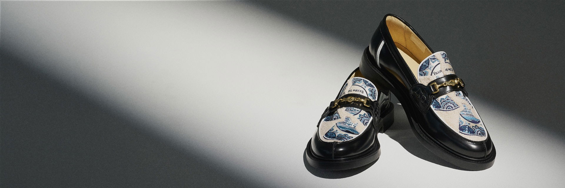 Schwarze Loafer mit goldener Schnalle und cremefarbenem Stoff mit blauem, floralem Muster, auf hellgrauem Untergrund. Schwarze Loafer mit goldener Schnalle und cremefarbenem Stoff mit blauem, floralem Muster, auf hellgrauem Untergrund.