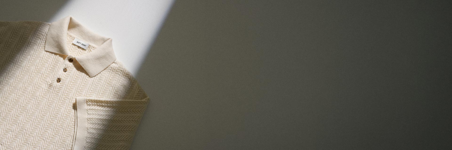 Beige Strickpolo mit Fischgrätmuster, kurzen Ärmeln und drei Knöpfen, liegend auf weißem und grauem Hintergrund. Beige Strickpolo mit Fischgrätmuster, kurzen Ärmeln und drei Knöpfen, liegend auf weißem und grauem Hintergrund.