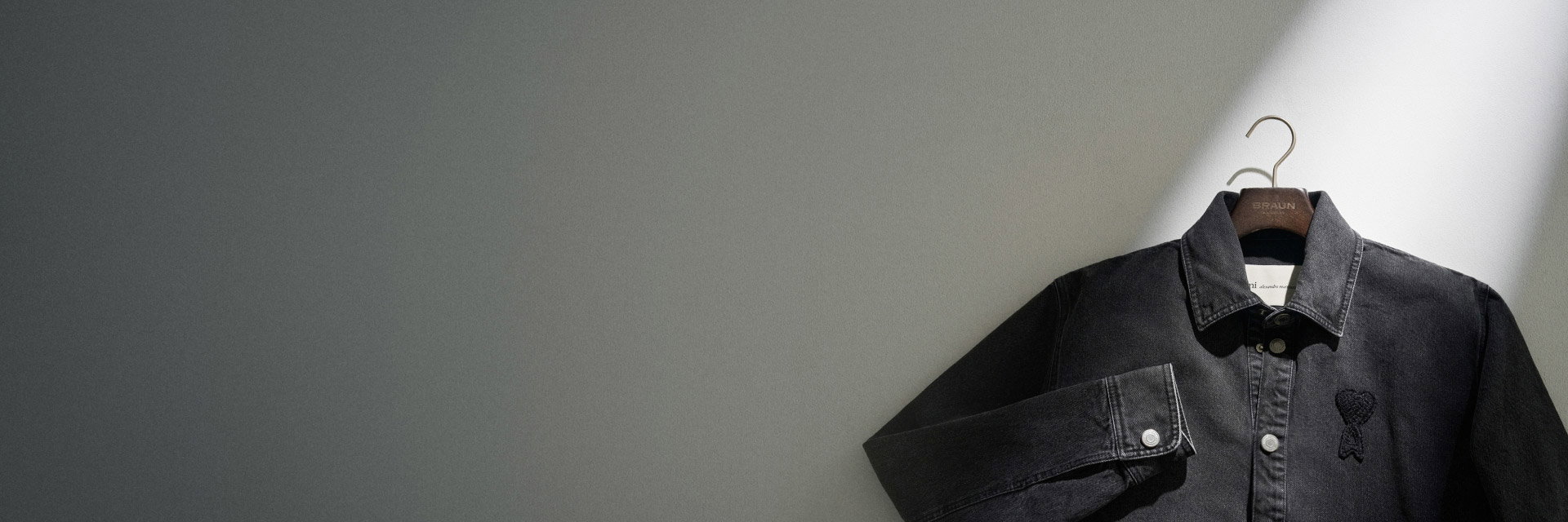 Schwarze Jeansjacke mit Kragen und Knöpfen, links Brustbereich mit schwarzem Schleifen-Logo, vor grauem Hintergrund auf Bügel. Schwarze Jeansjacke mit Kragen und Knöpfen, links Brustbereich mit schwarzem Schleifen-Logo, vor grauem Hintergrund auf Bügel.