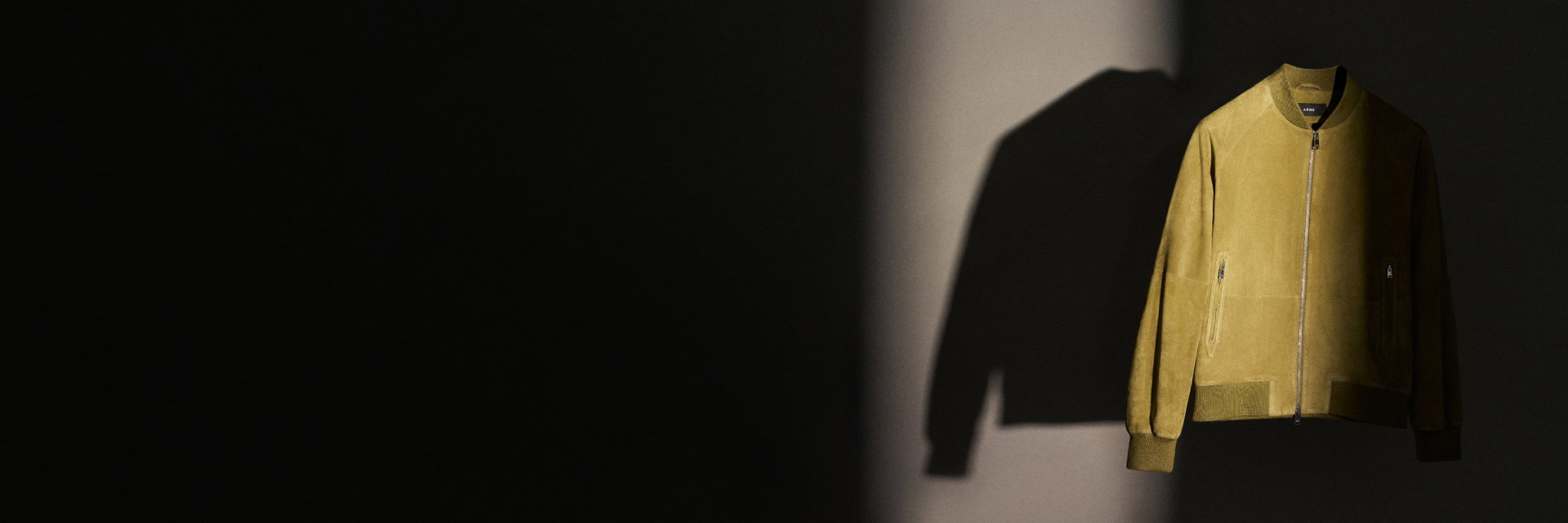 Olivgrüne Wildlederjacke mit Reißverschlüssen und gerippten Bündchen, vor dunklem Hintergrund hängend. Olivgrüne Wildlederjacke mit Reißverschlüssen und gerippten Bündchen, vor dunklem Hintergrund hängend.