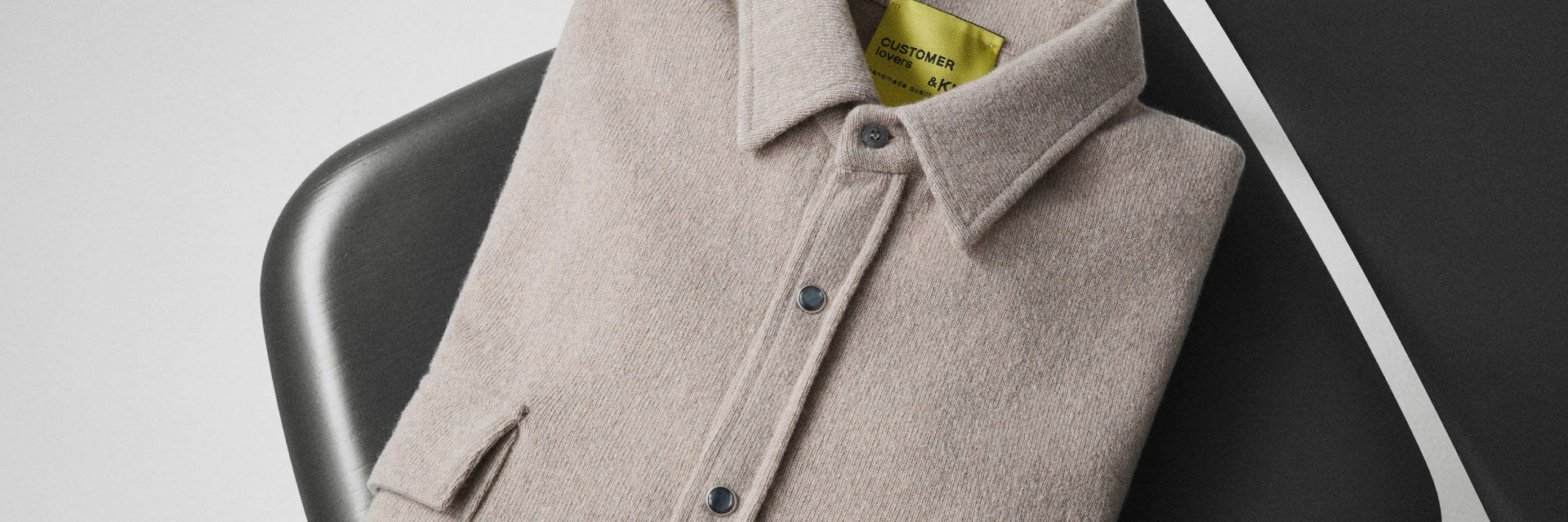 Beige Strickjacke mit Kragen und schwarzen Druckknöpfen, ordentlich gefaltet auf schwarzem Stuhl mit gelbem Etikett. Beige Strickjacke mit Kragen und schwarzen Druckknöpfen, ordentlich gefaltet auf schwarzem Stuhl mit gelbem Etikett.