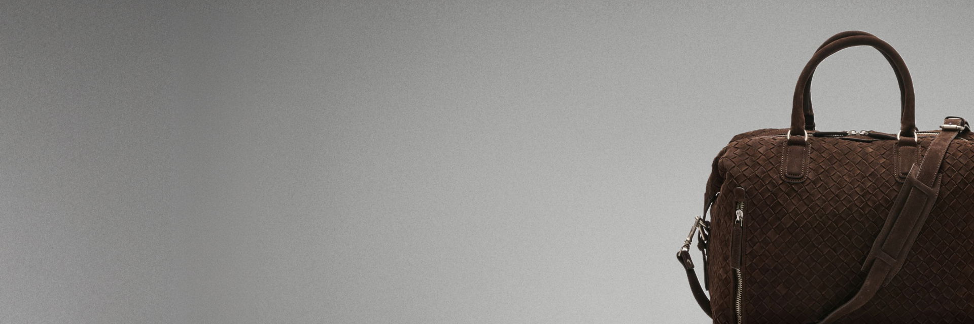 Braune, gewebte Lederreisetasche mit Griffen und verstellbarem Schultergurt vor grauem Hintergrund, rechts im Bild. Braune, gewebte Lederreisetasche mit Griffen und verstellbarem Schultergurt vor grauem Hintergrund, rechts im Bild.