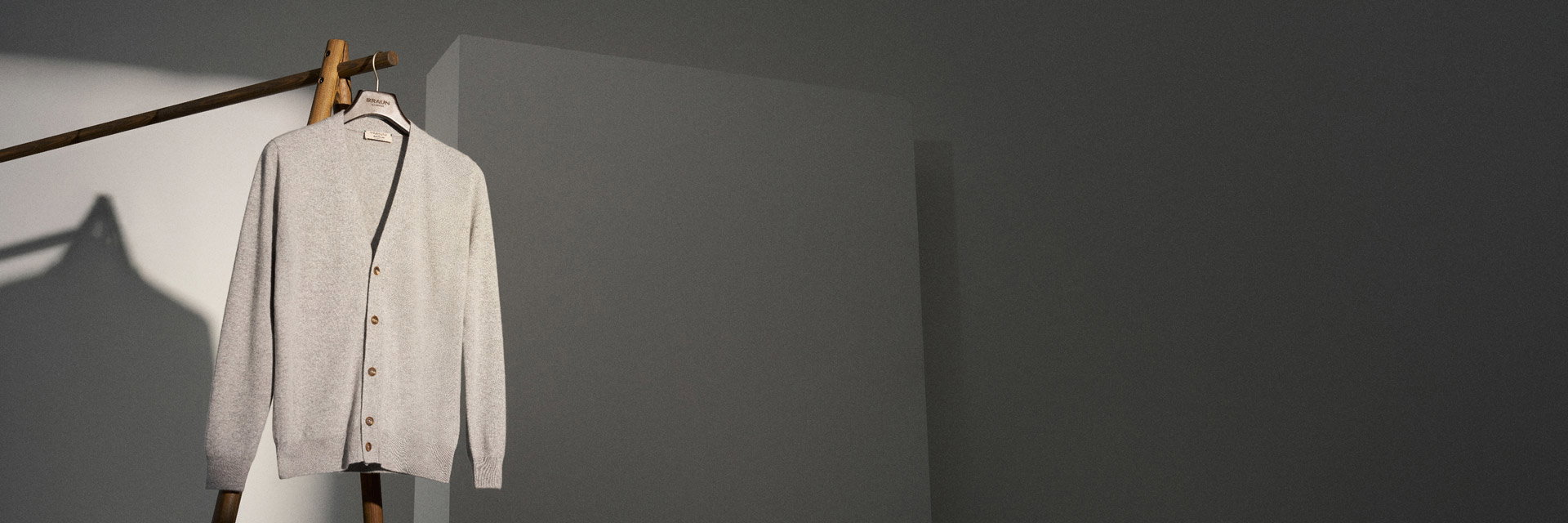 Beige Strickjacke mit Knöpfen an einem Holzkleiderbügel an einem Holzkleiderständer vor grauer Wand. Beige Strickjacke mit Knöpfen an einem Holzkleiderbügel an einem Holzkleiderständer vor grauer Wand.