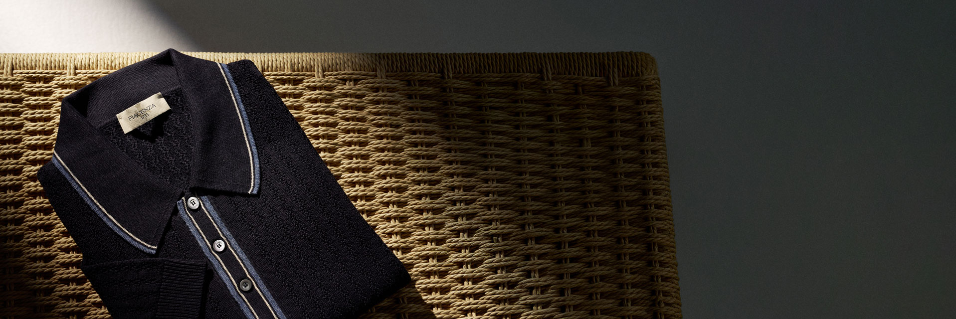 Dunkelblaues, gefaltetes Poloshirt mit weißen und blauen Streifen am Kragen und Knopfleiste auf geflochtenem Stuhl bei Sonnenlicht. Dunkelblaues, gefaltetes Poloshirt mit weißen und blauen Streifen am Kragen und Knopfleiste auf geflochtenem Stuhl bei Sonnenlicht.