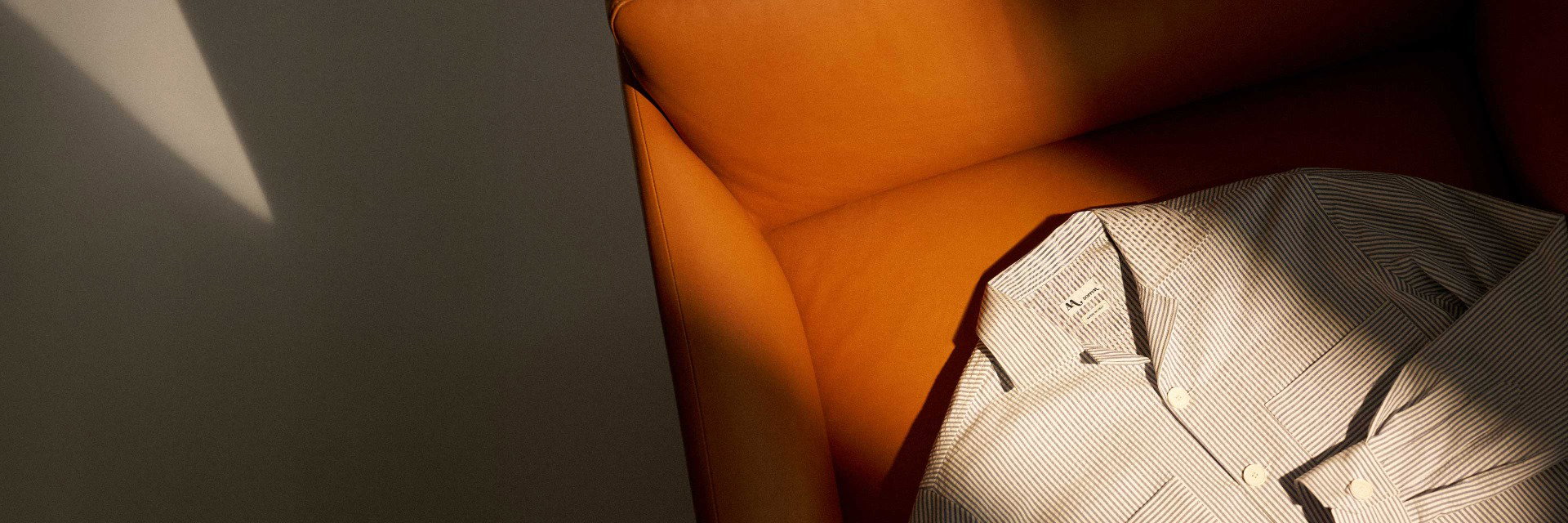 Beige-weiß gestreiftes Hemd liegt gefaltet auf einem orangefarbenen Ledersessel, Sonnenlicht fällt schräg ein. Beige-weiß gestreiftes Hemd liegt gefaltet auf einem orangefarbenen Ledersessel, Sonnenlicht fällt schräg ein.