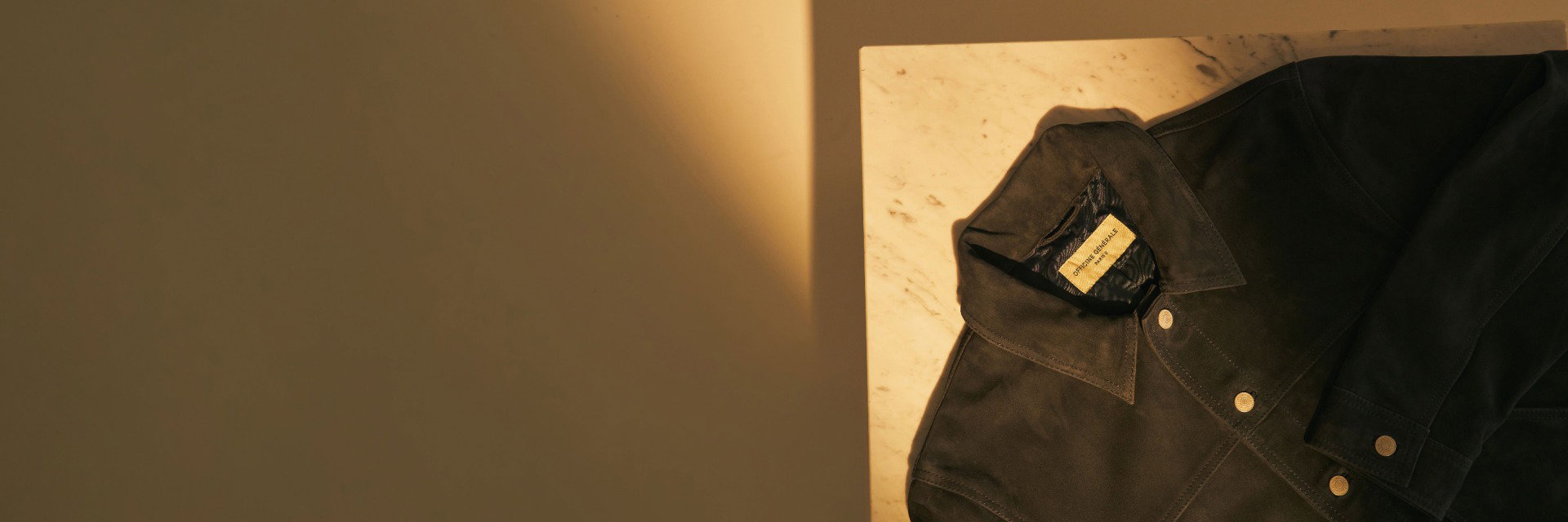 Elegante dunkelbraune Wildlederjacke mit Kragen und goldfarbenen Metallknöpfen auf hellem Marmoruntergrund, seitlich beleuchtet. Elegante dunkelbraune Wildlederjacke mit Kragen und goldfarbenen Metallknöpfen auf hellem Marmoruntergrund, seitlich beleuchtet.
