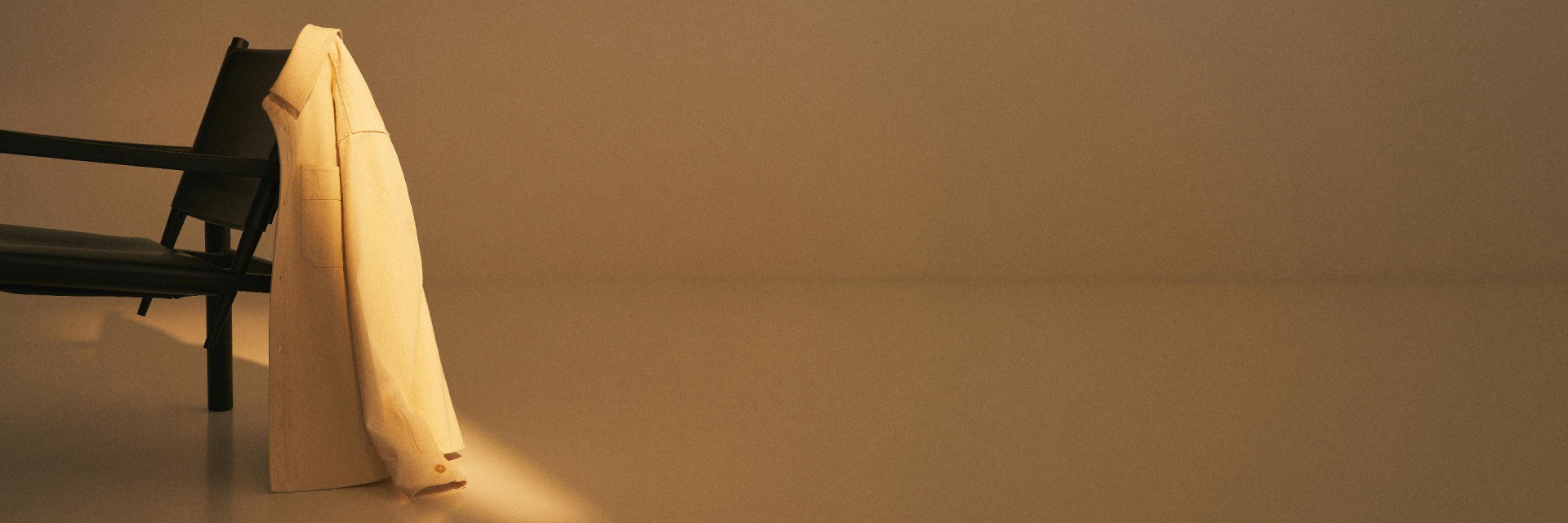Beige Jacke hängt über die Lehne eines schwarzen Stuhls vor einer hellbraunen schlichten Wand und Bodenfläche. Beige Jacke hängt über die Lehne eines schwarzen Stuhls vor einer hellbraunen schlichten Wand und Bodenfläche.