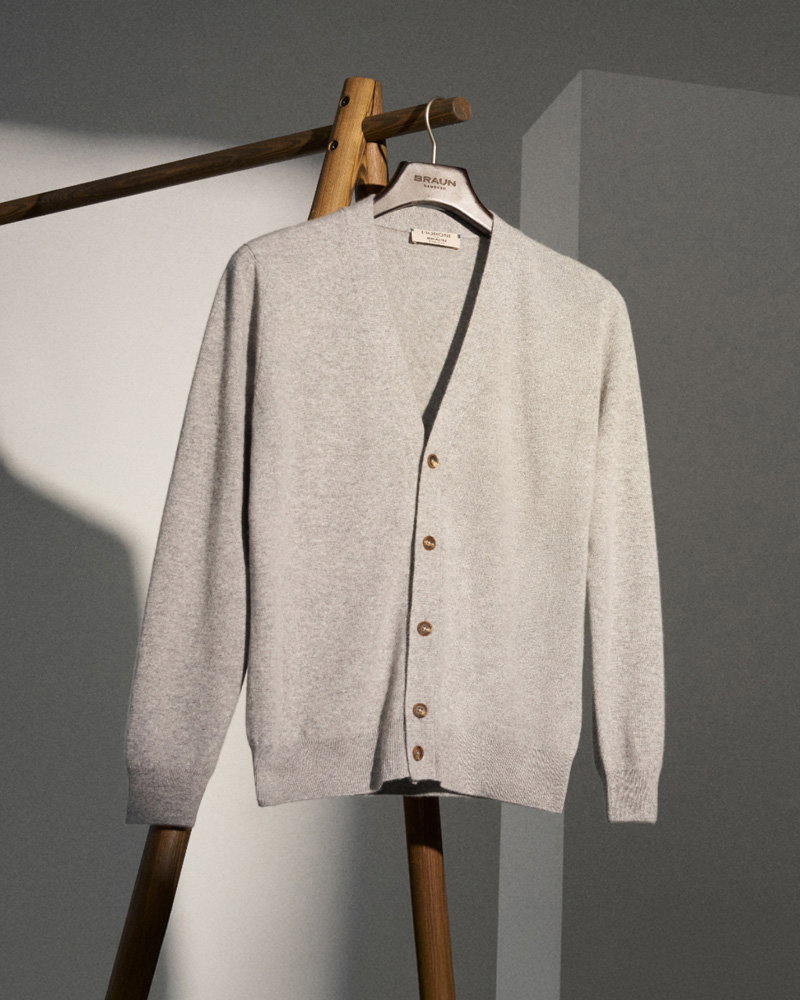 Light gray button-up cardigan hanging on a wooden hanger on a wooden clothing rack against a neutral wall.