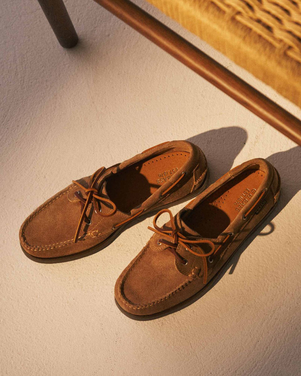 Braune Wildleder Bootsschuhe mit Schnürsenkeln liegen auf hellem Boden neben einem Stuhl mit Flechtgeflecht.